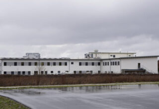 Ein modernes, zweistöckiges Gebäude mit flachem Dach und weißen Wänden. Das Gebäude der Analytik Institut Rietzler GmbH hat mehrere Fensterreihen und befindet sich auf einem großen, asphaltierten Gelände. Im Vordergrund ist eine nasse Asphaltfläche zu sehen, die den Regen reflektiert. Der Himmel ist bewölkt