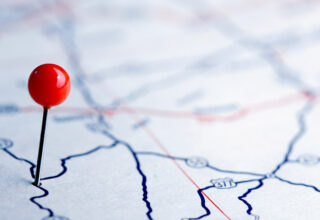 Eine Nahaufnahme einer Karte mit einer roten Stecknadel, die einen bestimmten Ort markiert. Die Karte zeigt verschiedene Straßen und Routen, dargestellt durch blaue und rote Linien