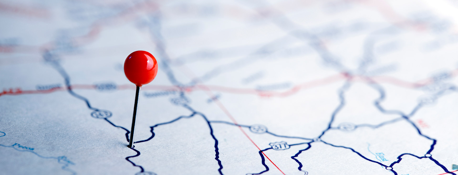Eine Nahaufnahme einer Karte mit einer roten Stecknadel, die einen bestimmten Ort markiert. Die Karte zeigt verschiedene Straßen und Routen, dargestellt durch blaue und rote Linien